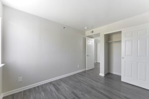 Interior unit bedroom, neutral toned walls, wood floors, sliding closet door, window parallel to bedroom door.